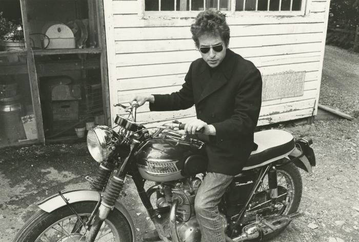 The photograph is in black and white. Bob Dylan on a motorcycle, named Triumph in the title, wearing a black coat, jeans, and sunglasses.